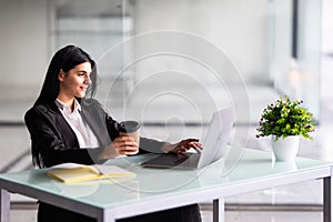 Attractive woman sitting at desk in office, working with laptop holding document, having takeaway coffee
