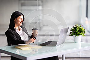 Attractive woman sitting at desk in office, working with laptop holding document, having takeaway coffee