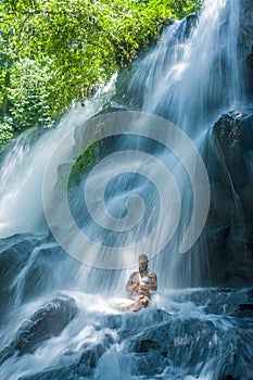 Attractive woman sitting at rock in yoga pose for spiritual relaxation serenity and meditation at stunning beautiful waterfall and