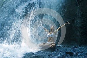 Attractive woman sitting at rock in yoga pose for spiritual relaxation serenity and meditation at stunning beautiful waterfall and