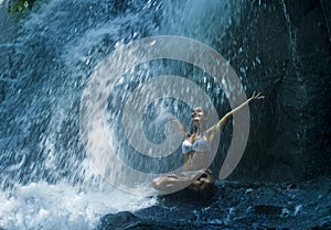 Attractive woman sitting at rock in yoga pose for spiritual relaxation serenity and meditation at stunning beautiful waterfall and