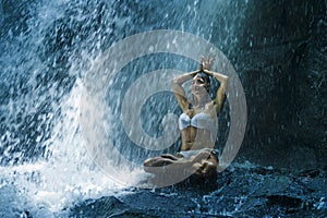 Attractive woman sitting at rock in yoga pose for spiritual relaxation serenity and meditation at stunning beautiful waterfall and