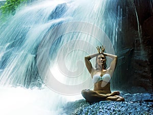 Attractive woman sitting at rock in yoga pose for spiritual relaxation serenity and meditation at stunning beautiful waterfall and