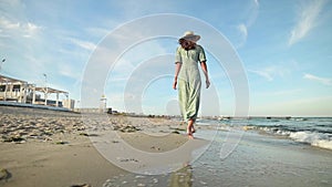 Attractive woman in slow motion walking barefoot along the beach in the early morning. Tourist in a light green dress on