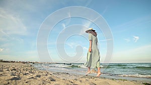 Attractive woman in slow motion walking barefoot along the beach in the early morning. Tourist in a light green dress on