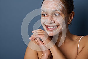 Attractive woman smiles with beautiful toothy smile, posing against gray background with facial mask, removing make-up and