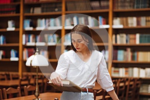 Attractive woman standing with a book in hand in a public library reads with a serious face on the background of shelves with