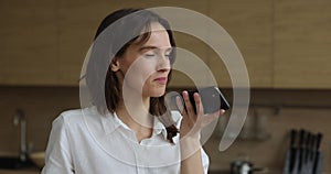 Attractive woman standing in kitchen holds smartphone talks on speakerphone