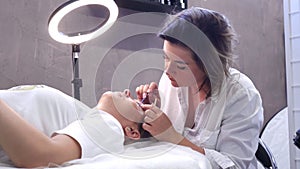 Attractive woman stylist, cosmetologist in white coat builds up, laminates eyelashes of client lying in front of her.