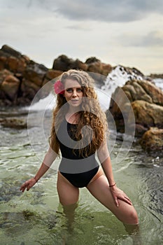 Attractive woman in swimsuit emerges from sea at dusk, red flower adorns her hair. Effortless beauty, serene beach