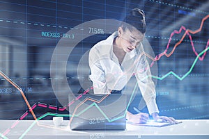 Attractive woman using laptop while leaning on desk creative business graph with index and grid on blurry office interior