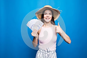 Attractive woman wear in straw hat showing banknotes of 100 usd, thumb-up, isolated over blue background