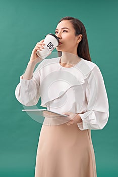 Attractive woman wearing shirt holding tablet and drinking takeaway coffee standing isolated over turquiose background