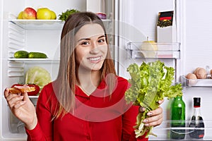 Attractive woman wears fashionable red blouse, stands near opened fridge, holds lettuce and sandwhich, going to prepare delicious