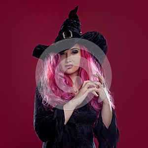 Attractive woman in witches hat and costume with red hair performs magic on pink background. Halloween, horror theme.