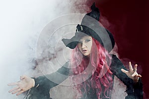 Attractive woman in witches hat with red hair performs magic. Smoke and witchcraft