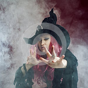 Attractive woman in witches hat with red hair performs magic. Smoke and witchcraft