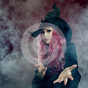 Attractive woman in witches hat with red hair performs magic. Smoke and witchcraft