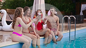 Attractive women in bikini clinking glasses with cocktails sitting by the swimming pool and flirting with fitted guy in