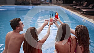 Attractive women in bikini clinking glasses with cocktails sitting by the swimming pool and flirting with fitted guy in