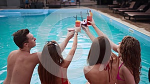 Attractive women in bikini clinking glasses with cocktails sitting by the swimming pool and flirting with fitted guy in