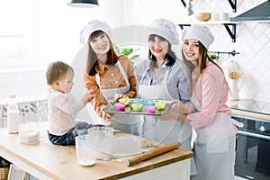 Attractive women in white aprons baking together, holding metal baking tray with colourful silicone forms for cupcakes