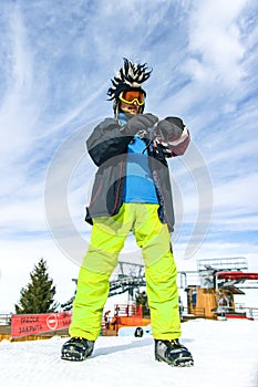Attractive youg man tourist in funny mohawk hat, green pants, ski glasses mask and black mittens on a background of blue