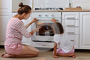 Attractive young adult mother wearing striped shirt and shorts sitting on floor near gas-stove with her daughter, female pointing