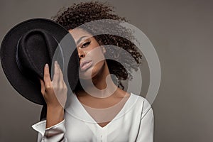 Attractive young african american woman in white shirt holding black hat over half of her face
