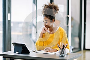 Attractive young African student studying at the college library, sitting at the desk, using a laptop computer, tablet and