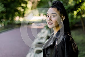 Attractive young Asian brunette woman in black leather jacket, relaxing on bench in the park outdoor. Close up of