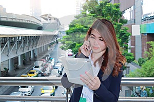 Attractive young Asian business woman talking on phone and looking document files in her hands at outside office background.