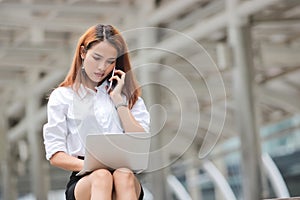 Attractive young Asian business woman using laptop and mobile smart phone for job at outside office.