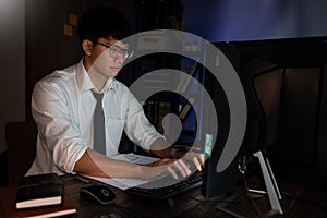 Attractive young asian businessman concentrated working until late overtime on publication at his desk in dark modern office at