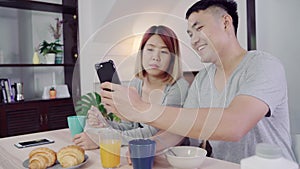 Attractive young Asian couple distracted at table with newspaper and cell phone while eating breakfast.