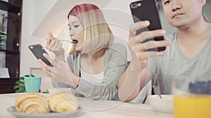 Attractive young Asian couple distracted at table with newspaper and cell phone while eating breakfast.