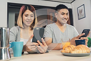 Attractive young Asian couple distracted at table with newspaper and cell phone while eating breakfast. Excited young Asian couple