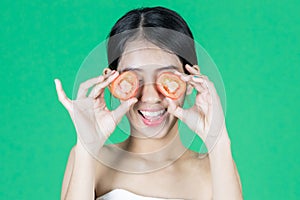 Attractive young Asian woman covering her eyes with tomato slice over green isolated background. Healthy and beauty skin care