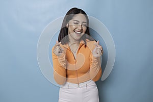 Attractive young Asian woman feels happy and romantic shapes heart gesture expresses tender feelings wears casual orange top