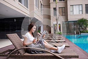 Attractive young Asian woman using tablet and drinking water near swimming pool. Relaxation concept