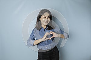 An attractive young Asian woman wearing a blue shirt feels happy and a romantic shapes heart gesture expresses tender feelings