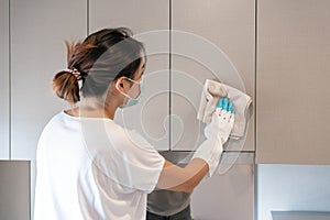 Attractive young Asian woman wearing rubber protective white gloves cleaning a surface of kitchen wall cabinet with rag. Home,