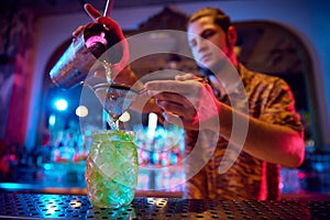 Attractive young bartender gently pouring refreshing mojito from shaker into cocktail glass on bar counter at night club