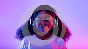 Attractive young black lady listening to music in wireless headphones, smiling with closed eyes in neon light, panorama