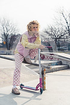 Attractive young blond Caucasian woman posing with a scooter on the skateboard ground, smiling and looking away, bright