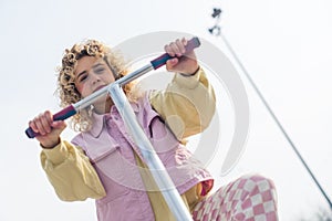 Attractive young blond hipster woman keeping her hands on the pink scooter, smiling and looking at the camera copy space