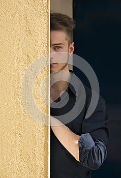 Attractive young blond man hiding half face behind wall