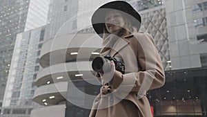 Attractive young blond woman with a camera in her hands in the city street. Action. Elegant female in brown coat and