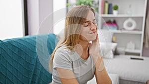 Attractive young blonde adult woman sitting on living room sofa at home, suffering in agony from severe teeth pain - oral issue