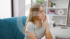 Attractive young blonde adult woman sitting on living room sofa at home, suffering in agony from severe teeth pain - oral issue